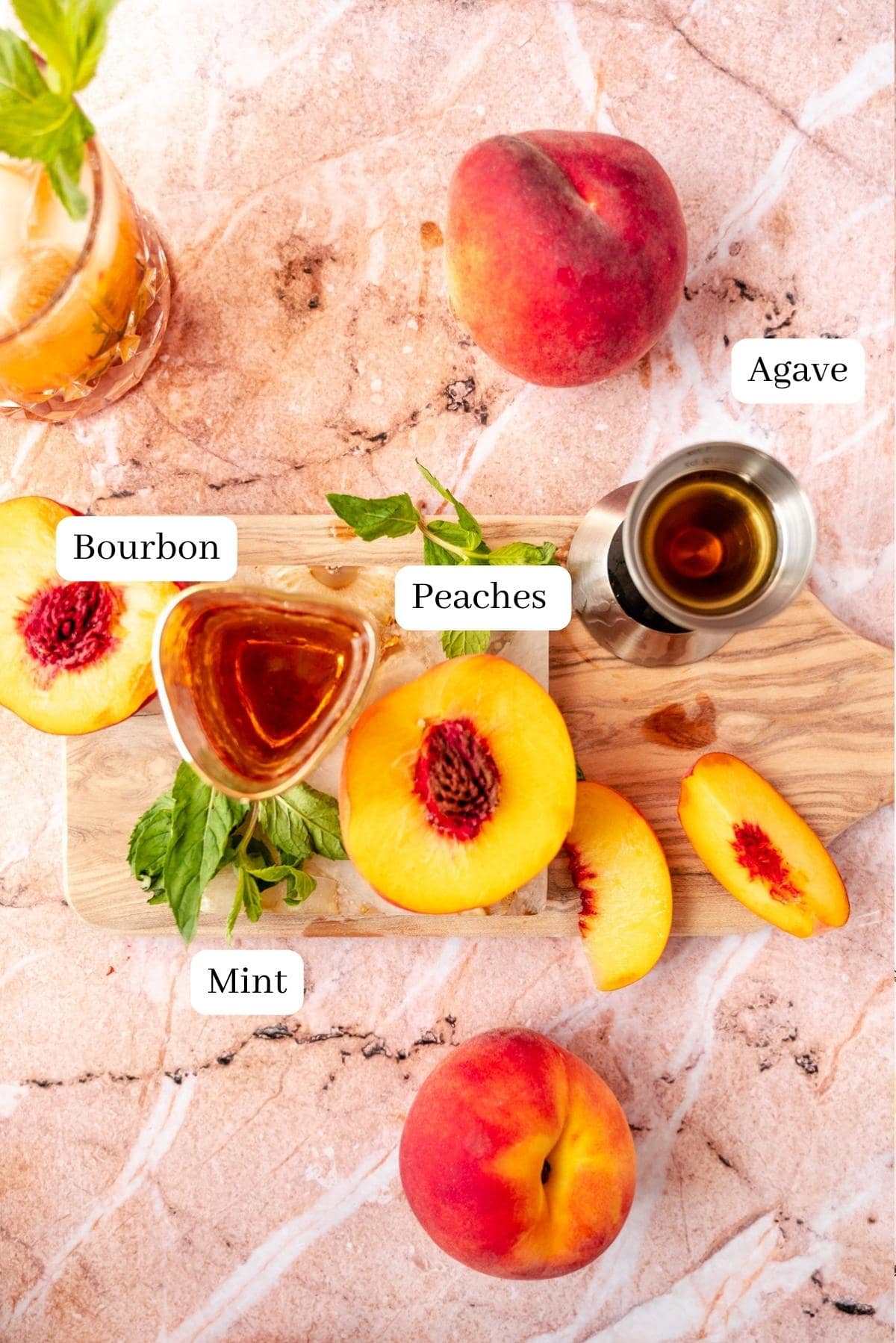 The ingredients for a peach bourbon smash on a pink marble table.