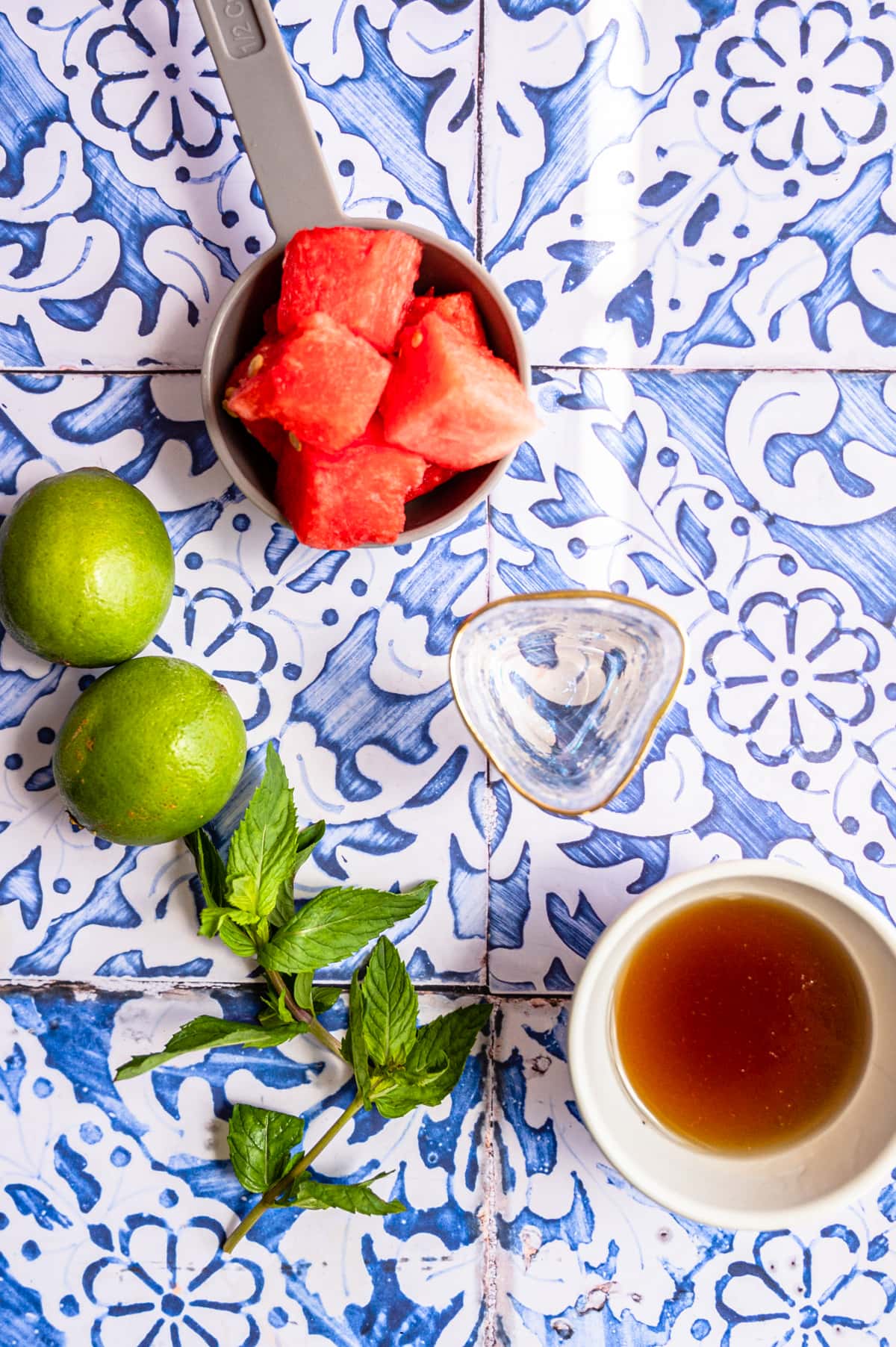 A shot glass with vodka, cubed watermelon, limes,  mint and agave on a blue tile table.