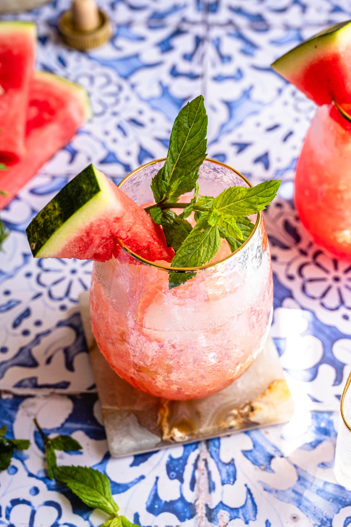 A watermelon smash cocktail on a blue tile table with a large sprig of mint and a slice of watermelon garnished on the glass.