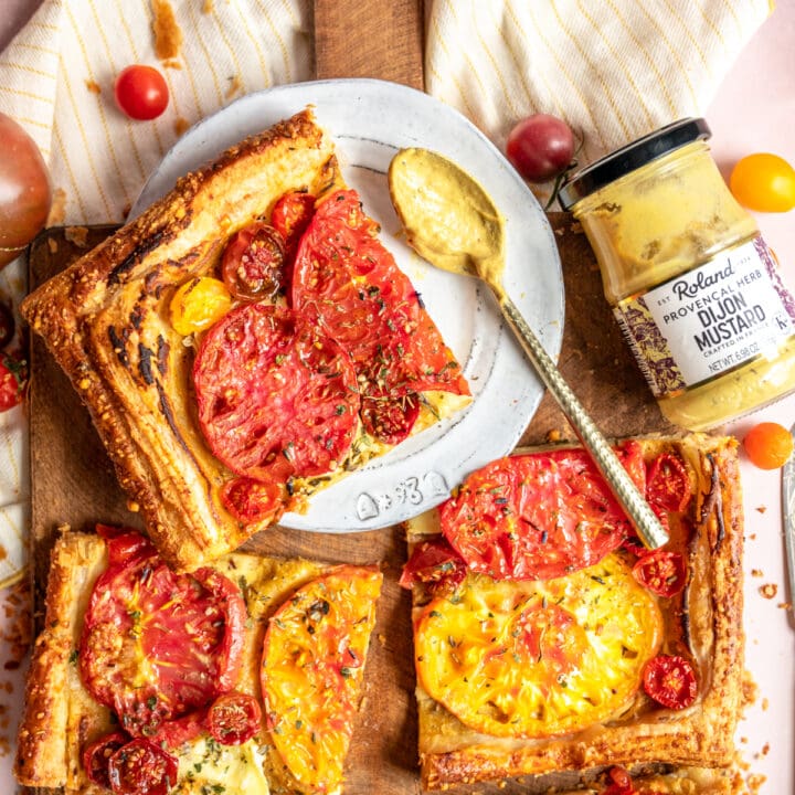 A sliced tomato tart on a wood cutting board with one grey dessert plate with a slice sitting on top. A jar of Roland Foods Mustard is off to the side.