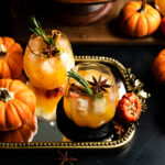 Two pumpkin glasses and a pumpkin punch bowl filled with pumpkin punch on a black tile table.