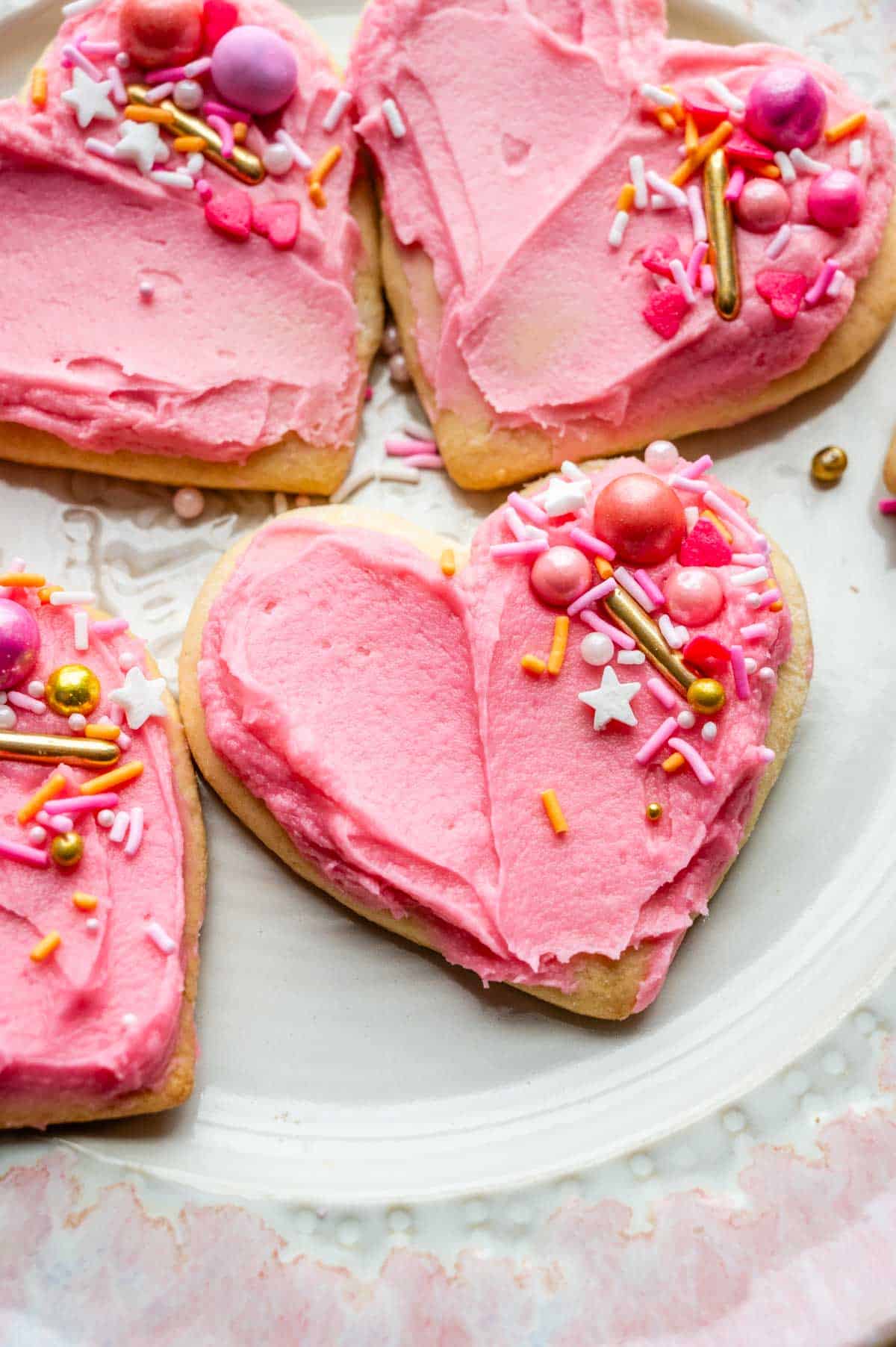 Pink frosted heart shaped sour cream cookies with sprinkles sitting on a plate with pink trim.