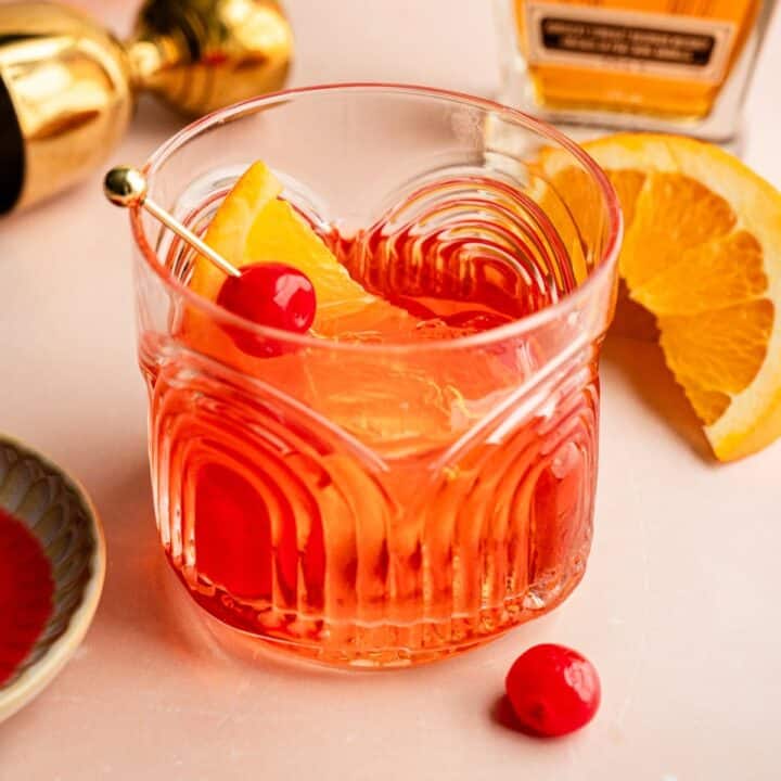 An aperol old fashioned garnished with an orange wedge and a cherry sitting on a pink marble table.
