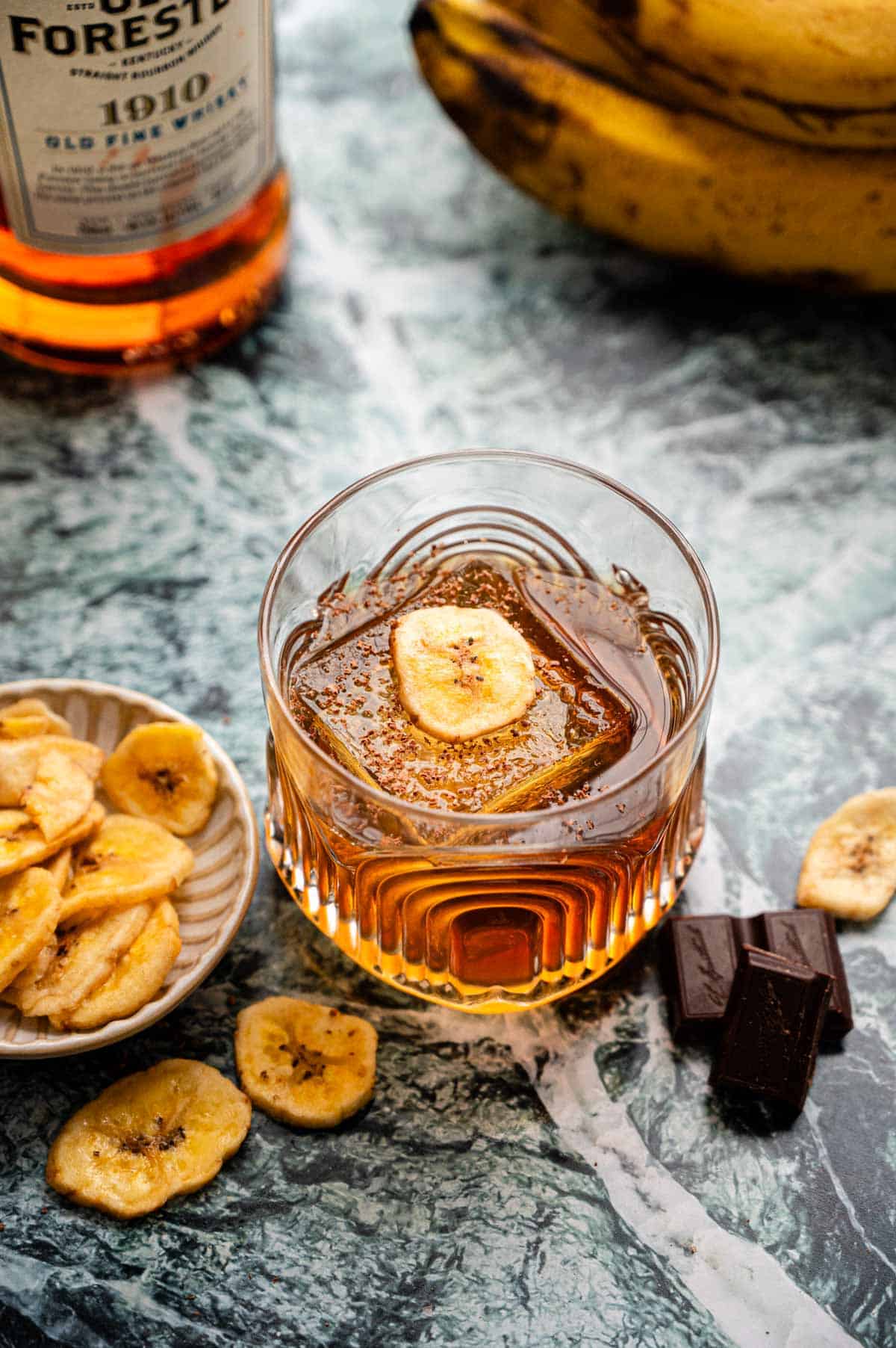 A rocks glass with a banana old fashioned  garnished with chocolate shavings and a banana chip, sitting on a green marble table.