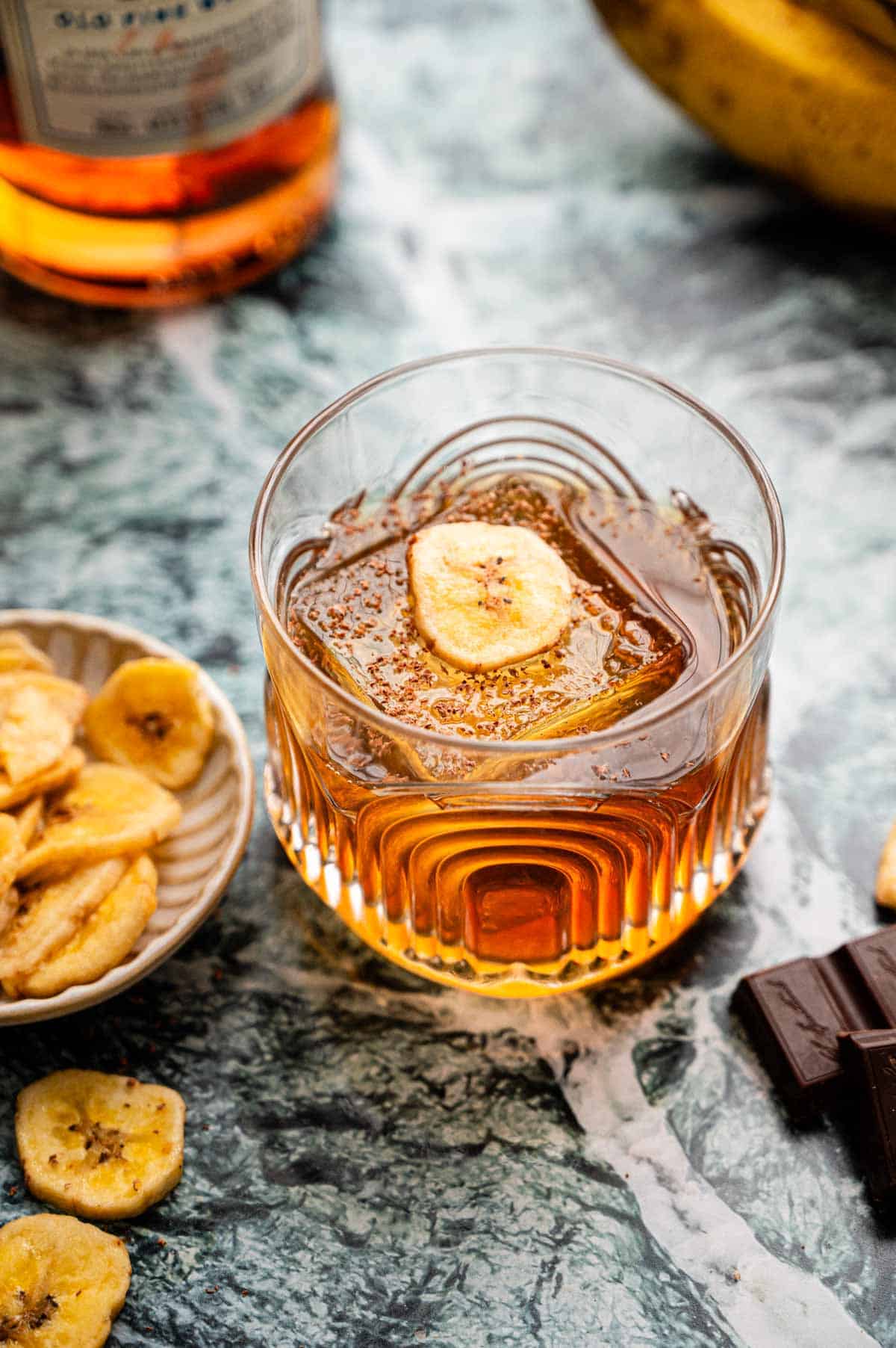 A rocks glass with a banana old fashioned garnished with chocolate shavings and a banana chip, sitting on a green marble table.