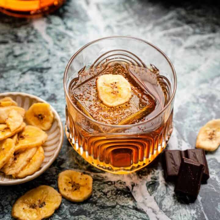 A rocks glass with a banana old fashioned garnished with chocolate shavings and a banana chip, sitting on a green marble table.
