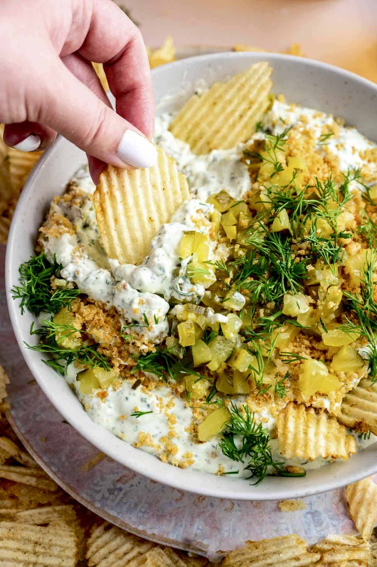 A bowl full of dip topped with bread crumbs, chopped, pickles, and dill with a hand scooping some dip with a chip.