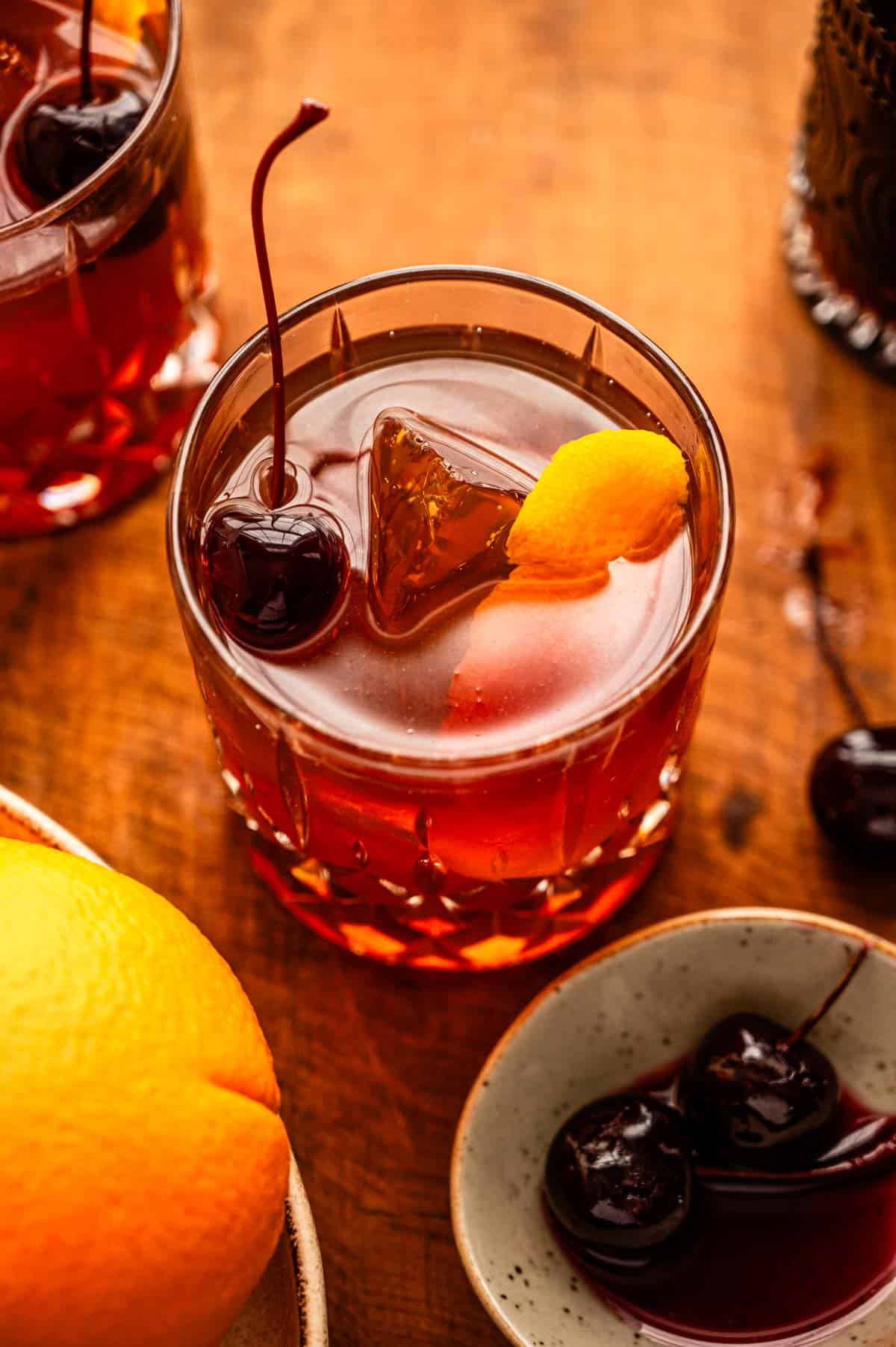 An overhead shot of an old fashioned with an orange peel and cheery in the glass. The glass is on a wood cutting board with a orange and cherries off to the side.