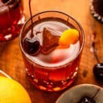 An overhead shot of an old fashioned with an orange peel and cheery in the glass. The glass is on a wood cutting board with a orange and cherries off to the side.