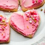 Pink frosted heart shaped sour cream cookies with sprinkles sitting on a plate with pink trim.