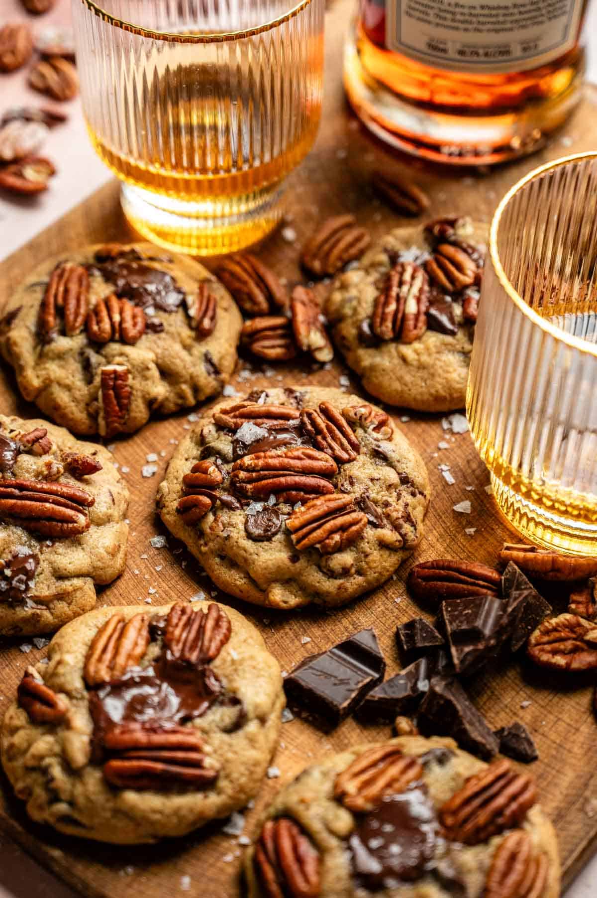 Bourbon pecan chocolate chip cookies on a wood cutting board with two glasses of bourbon and chocolate bar pieces surrounding the cookies.