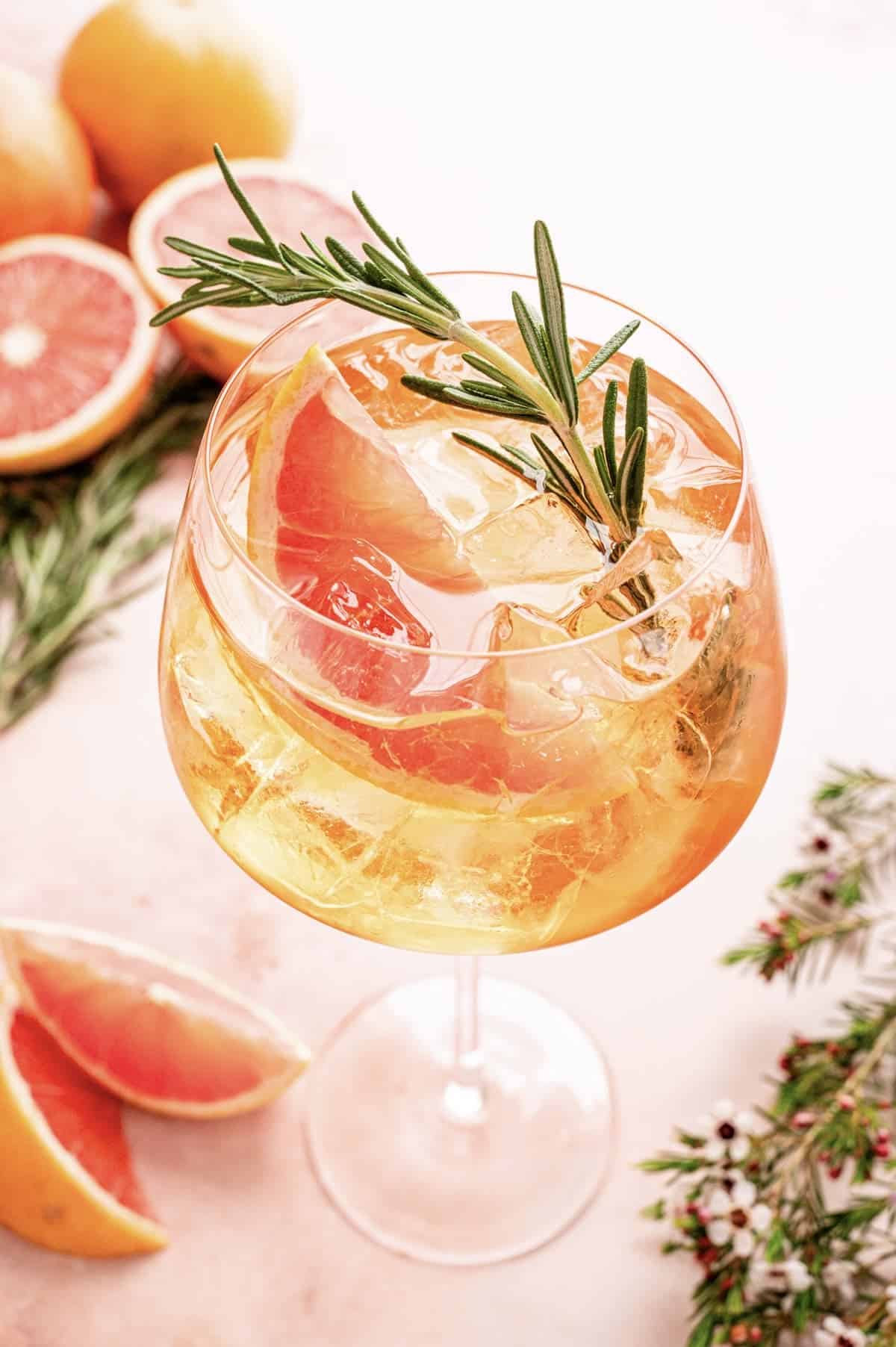 Grapefruit aperol spritz in a large wine glass and garnished with a grapefruit wedge and a rosemary sprig on a pink marble table.