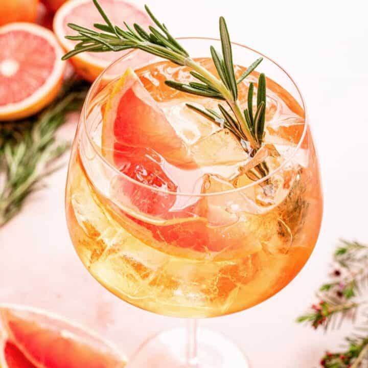 Grapefruit aperol spritz in a large wine glass and garnished with a grapefruit wedge and a rosemary sprig on a pink marble table.