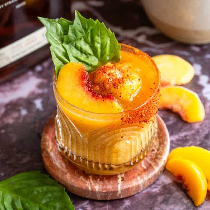 The peach bourbon frozen drink garnished with a peach slice, basil, and tan on a purple marble table.