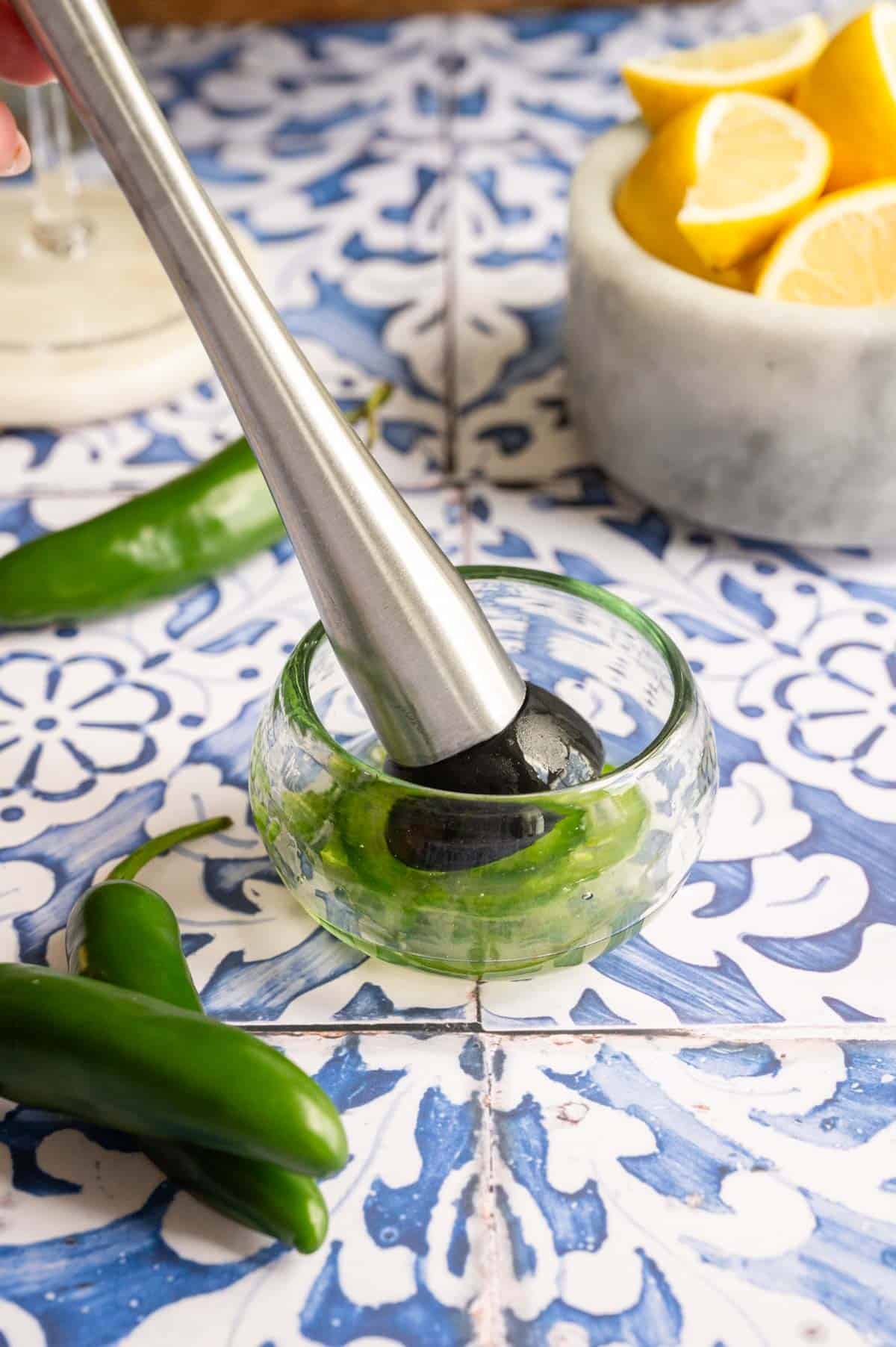 A small glass bowl with a muddler and jalapeño slices on a blue tile table.