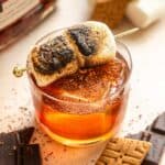 A small glass with the bourbon cocktail garnished with toasted marshmallows on a brown table.