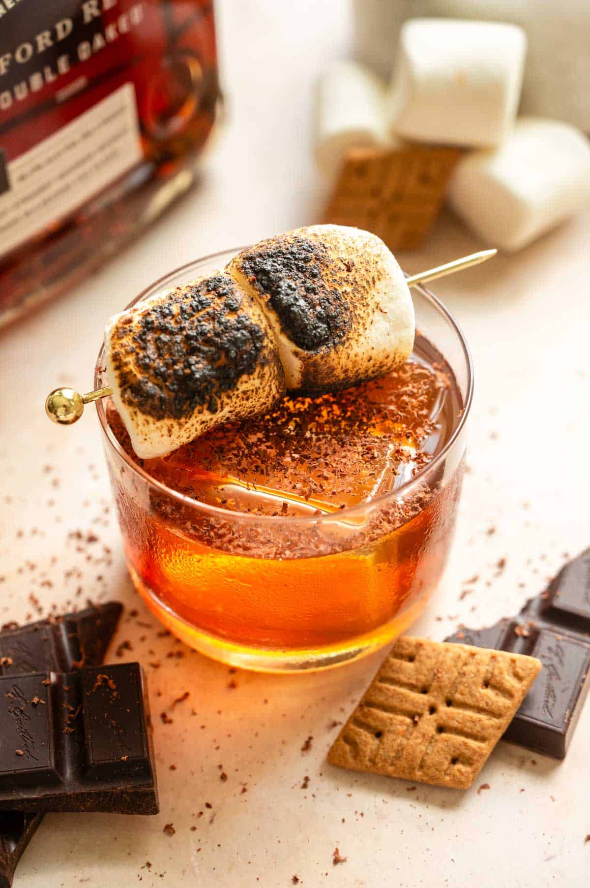 A small glass with the bourbon cocktail garnished with toasted marshmallows on a brown table.