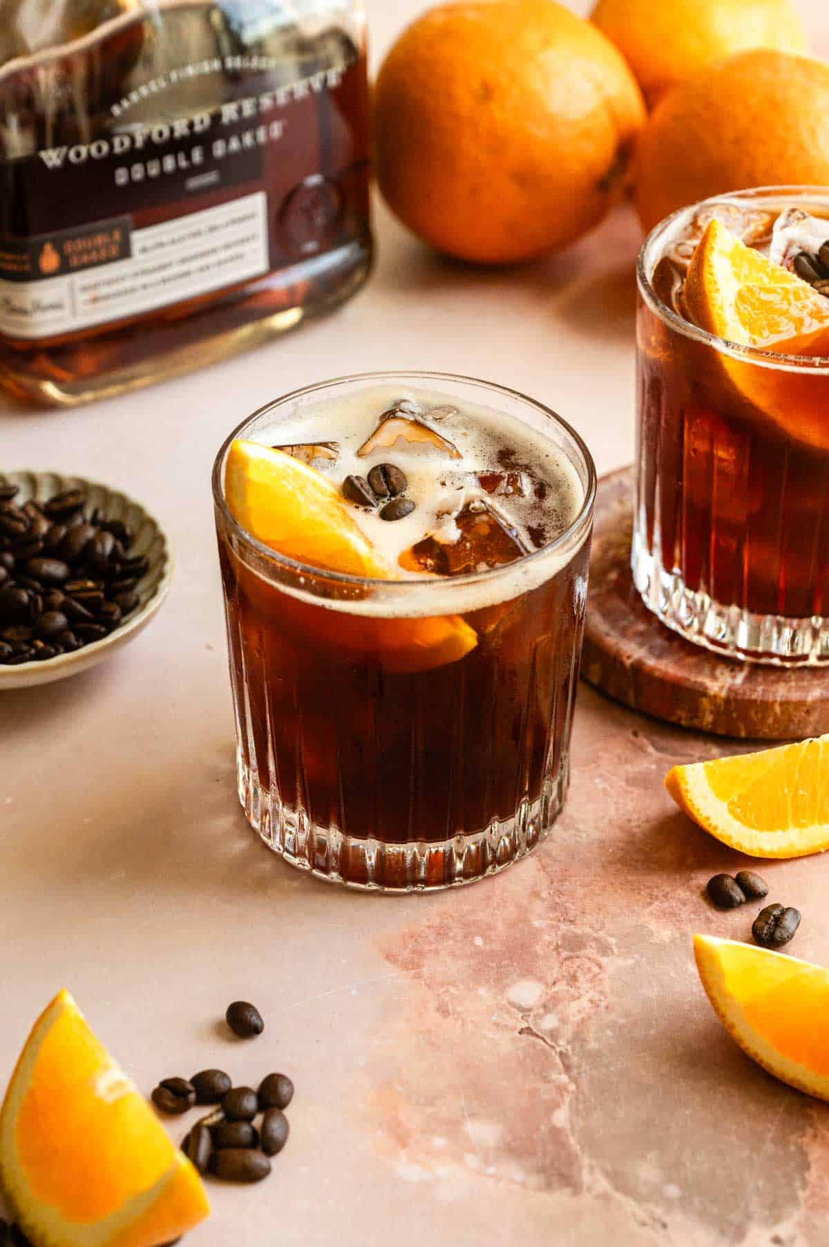 Two bourbon Carajillo cocktails in a rocks glasses and garnished with an orange slice on a pink marble table