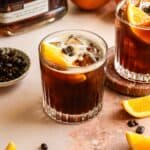 Two bourbon Carajillo cocktails in a rocks glasses and garnished with an orange slice on a pink marble table