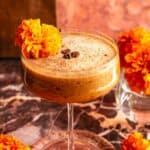 An espresso martini in a coupe glass and garnished with a marigold and espresso beans on a mauve marble table.