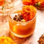 A small rocks glass filled with the cocktail on a orange marble table and garnished with star anise and a marigold.