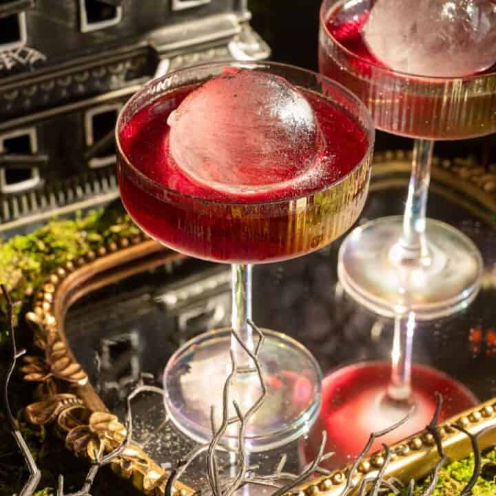 Two coupe glasses with a circle ice cube surrounded by a dark red sparkly cocktail.