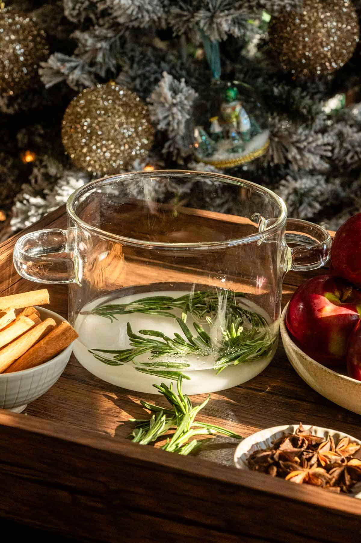 sugar, water, and rosemary in a glass pot ready to be cooked.