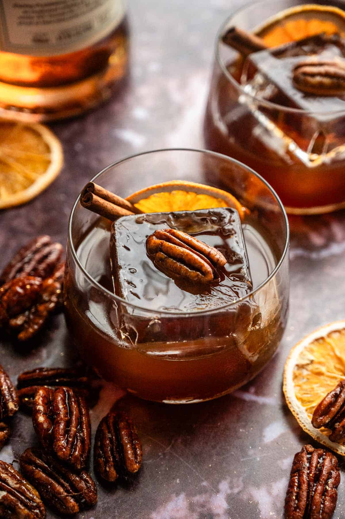 A candied pecan old fashioned garnished with an orange, cinnamon, stick, and a candied pecan on a purple marble table.