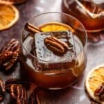 A candied pecan old fashioned garnished with an orange, cinnamon, stick, and a candied pecan on a purple marble table.