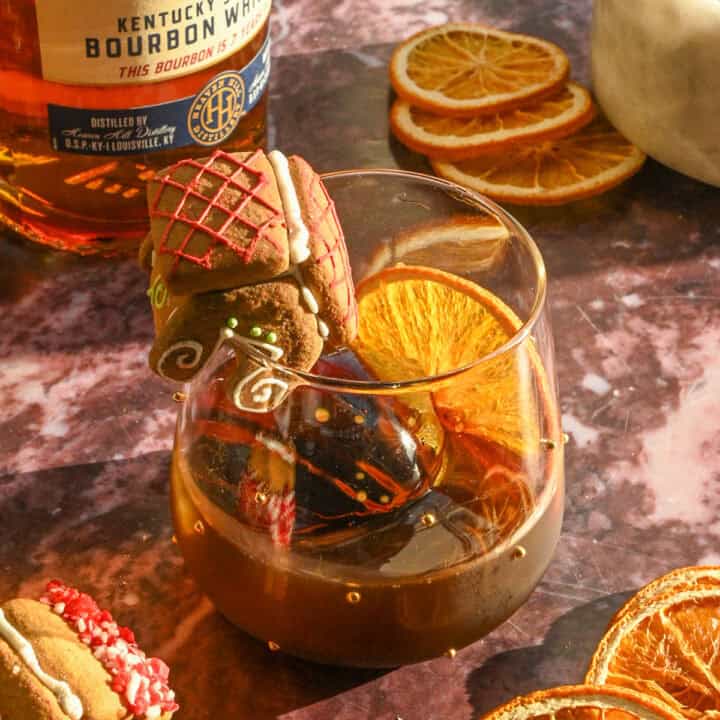 A gingerbread old fashioned garnished with and ornage slice and a little gingerbread house on a purple marble table.