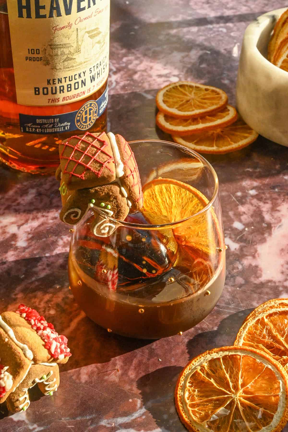 A gingerbread old fashioned garnished with and ornage slice and a little gingerbread house on a purple marble table.