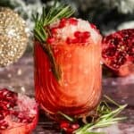 A pomegranate julep filled with fine crushed ice and garnished with rosemary on a purple marble table.