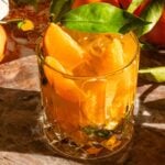 A mandarin smash in a rocks glass on a pink marble slab and garnished with a mandarin leaf.