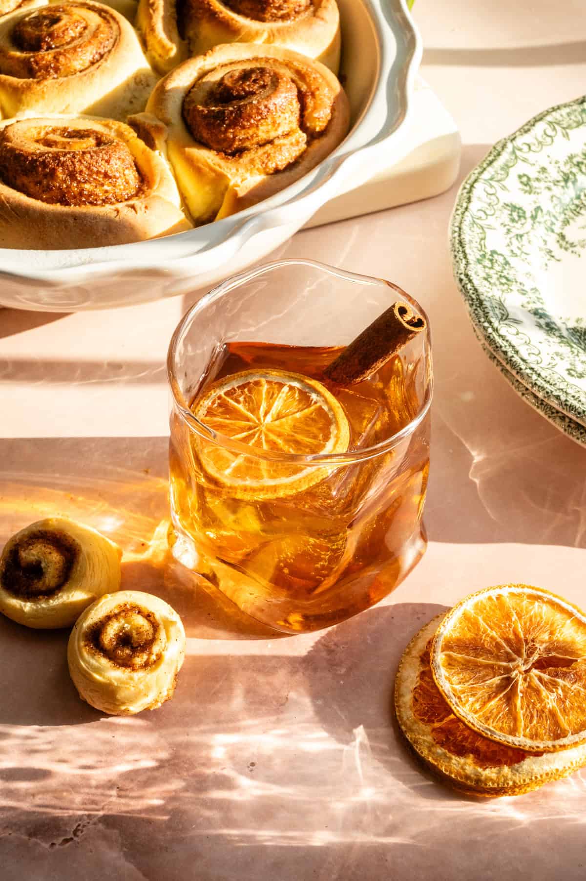 A maple cinnamon old fashioned in a slightly distorted glass and garnished with a dried orange slice and cinnamon stick.