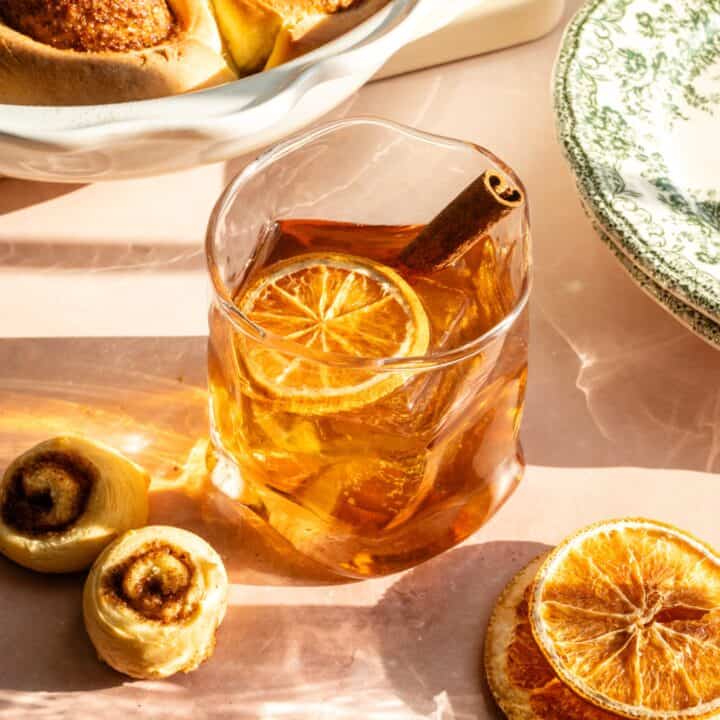 A maple cinnamon old fashioned in a slightly distorted glass and garnished with a dried orange slice and cinnamon stick.