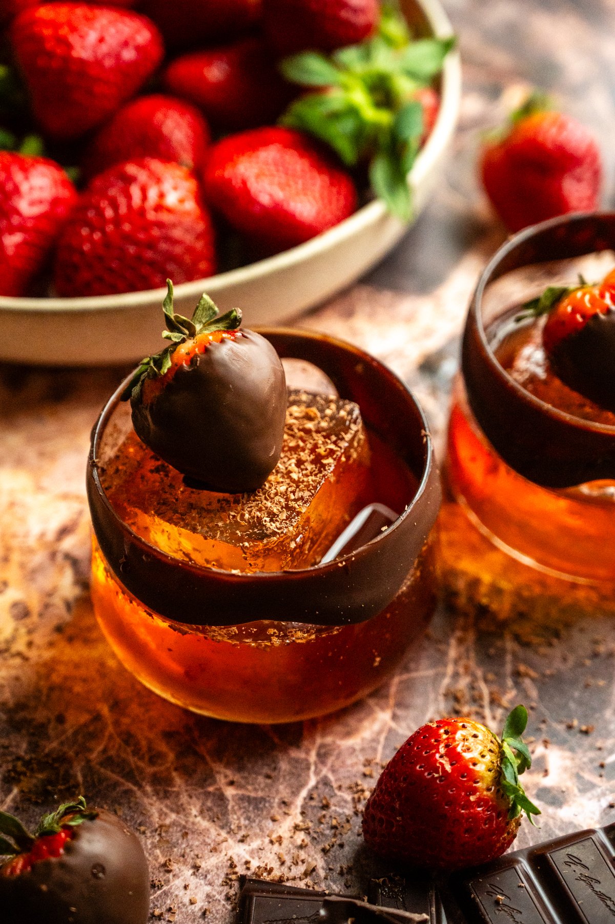 Two chocolate covered strawberry old fashioned on a pink marble table and garnished with a chocolate strawberry.