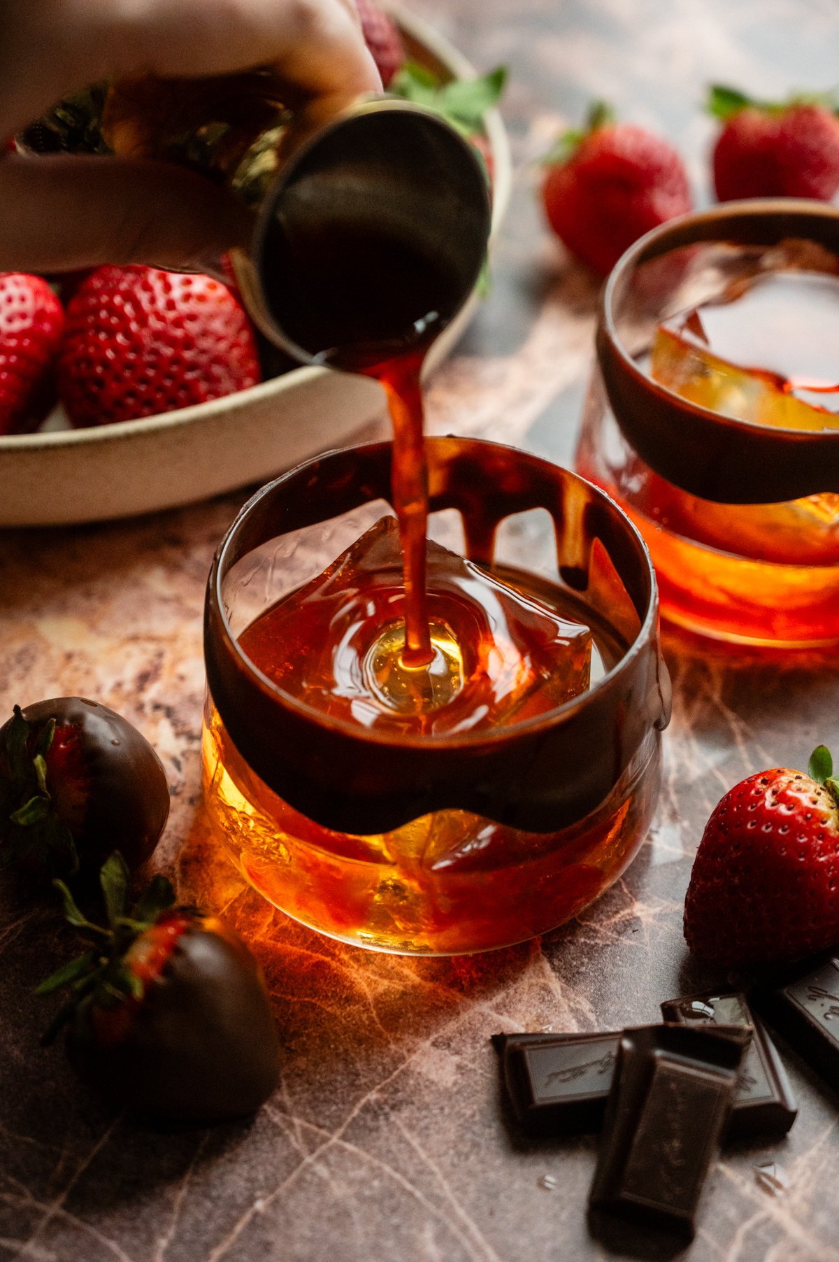 Strawberry syrup is being poured into a chocolate rimmed rocks glass with a large ice cube.