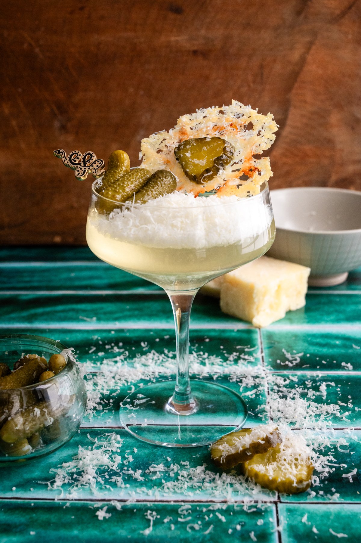 A parmesan and pickle martini on a green tile table with parmesan cheese almost filling the glass and its garnished with a parmesan crisp.