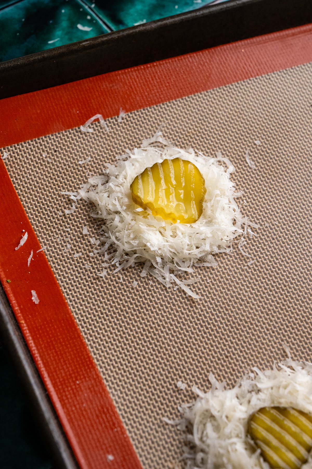 A spoonful of parmesan cheese with a pickle on top on a lined baking sheet.