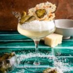 A parmesan and pickle martini on a green tile table with parmesan cheese almost filling the glass and its garnished with a parmesan crisp.