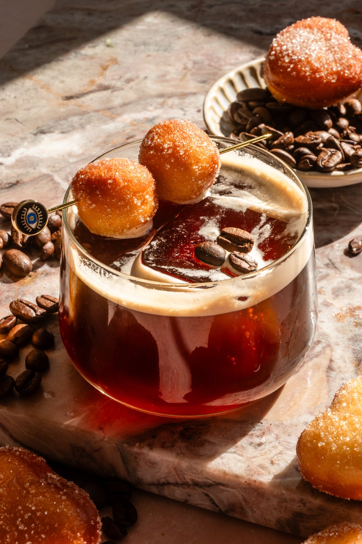 A vanilla espresso old fashioned with two mini donut holes on a pink marble table.