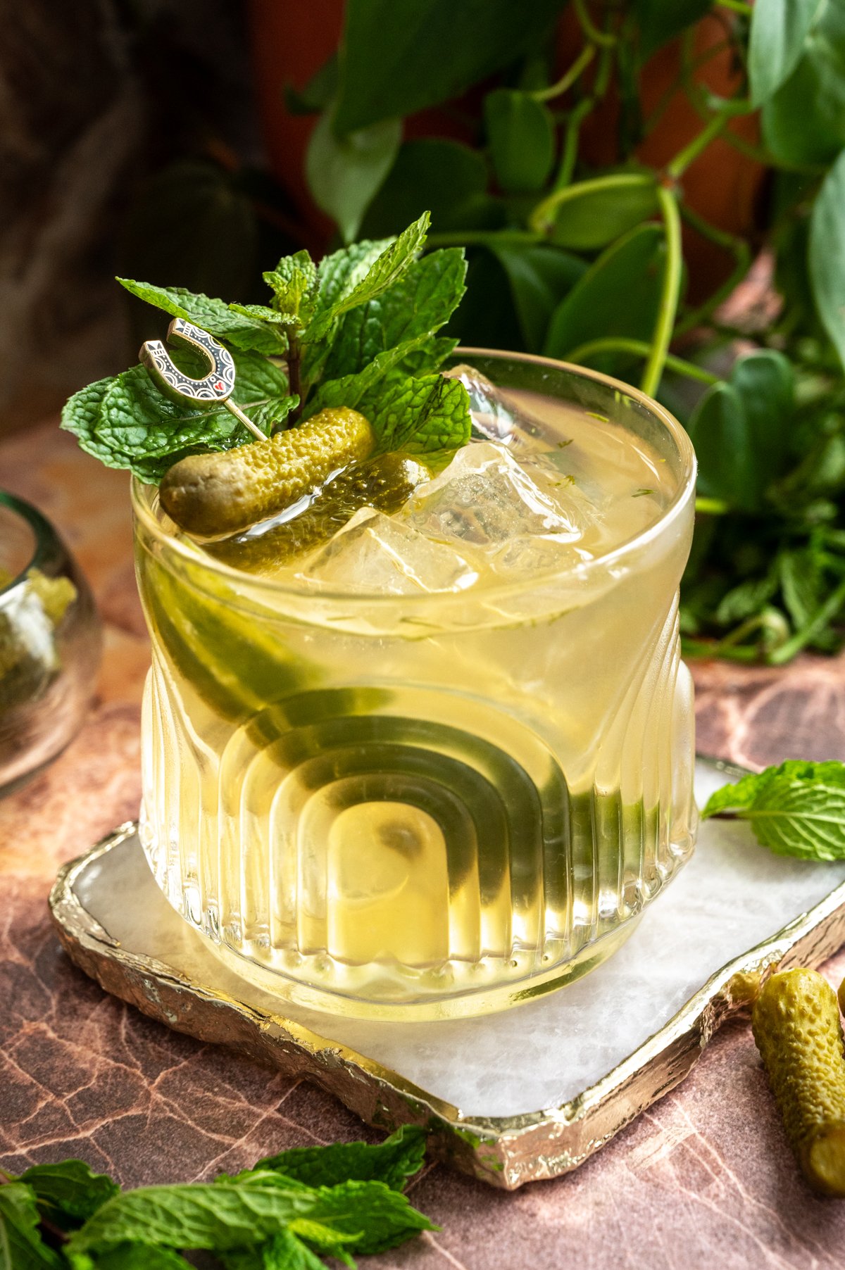 A rocks glass filled with the cocktail and garnished with a pickle pear, mint and mini pickles on a pink marble back ground.