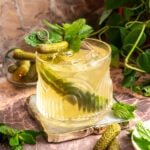 A rocks glass filled with the cocktail and garnished with a pickle pear, mint and mini pickles on a pink marble back ground.