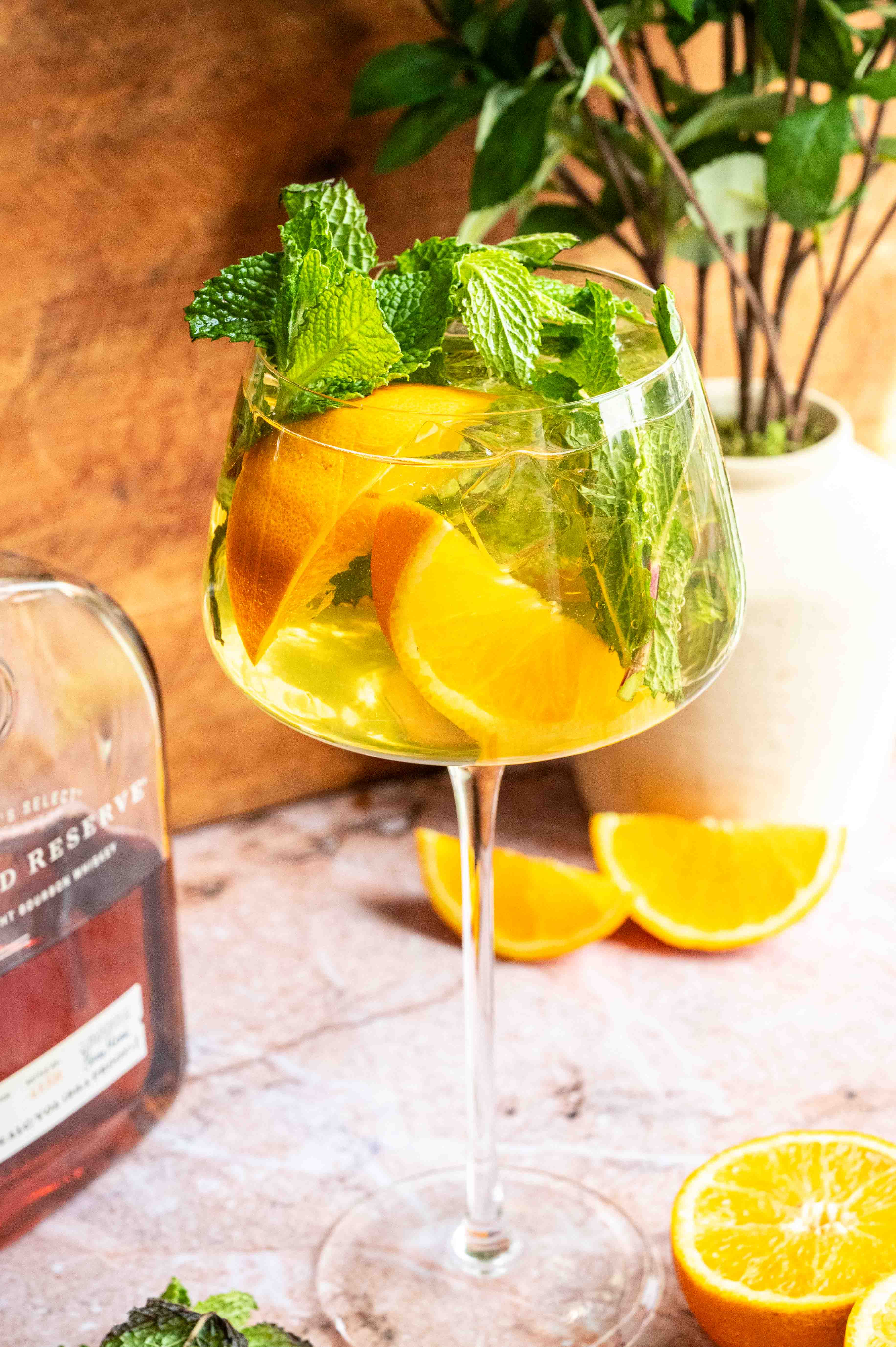 A long stemmed spritz glass filled with the cocktail and garnished with lots of mint and orange wedges on a pink marble table.