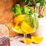 A long stemmed spritz glass filled with the cocktail and garnished with lots of mint and orange wedges on a pink marble table.