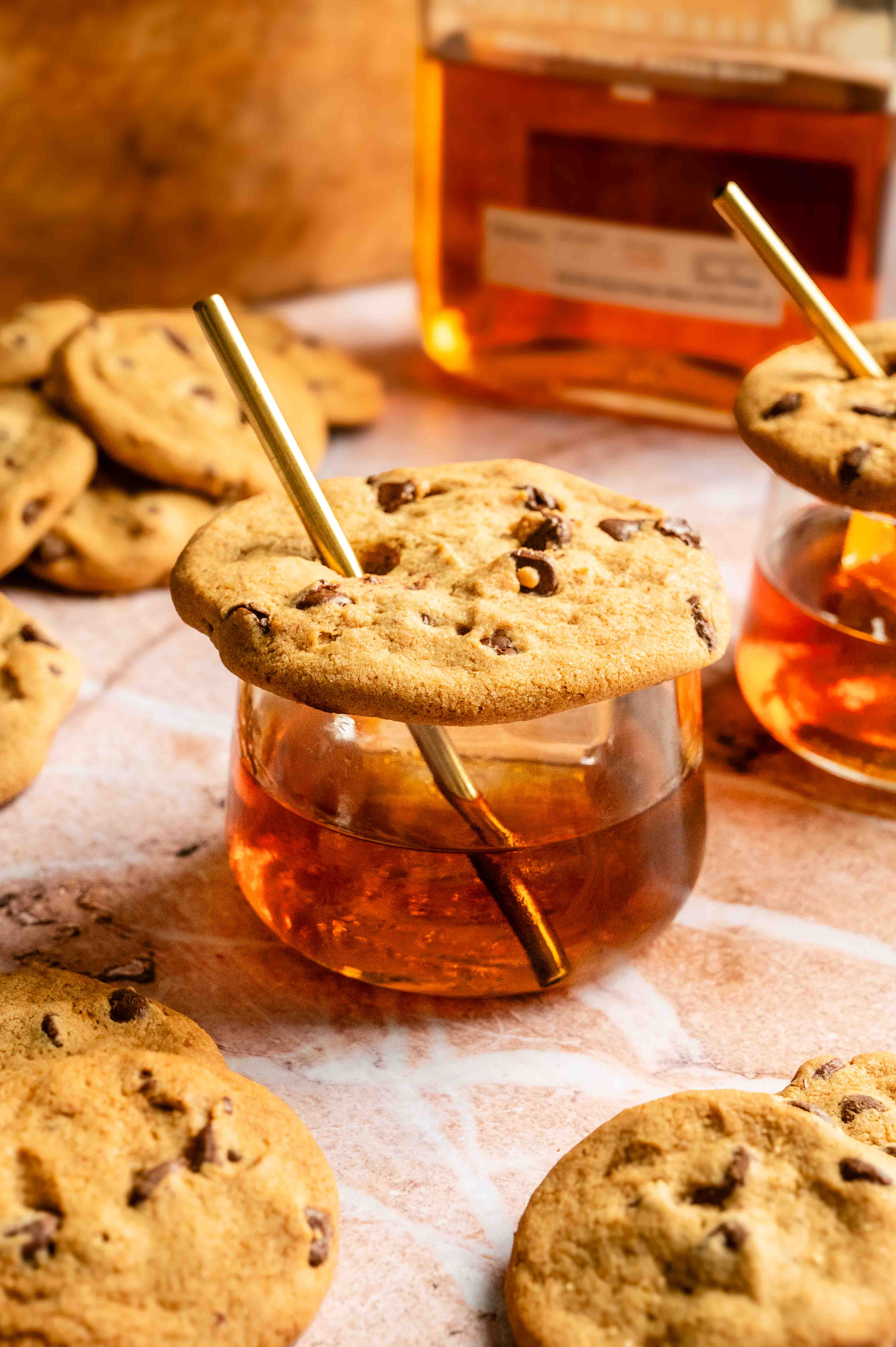 A small old fashioned glass filled with an chocolate chip cookie old fashioned and topped with a chocolate chip cookie with a straw through it.