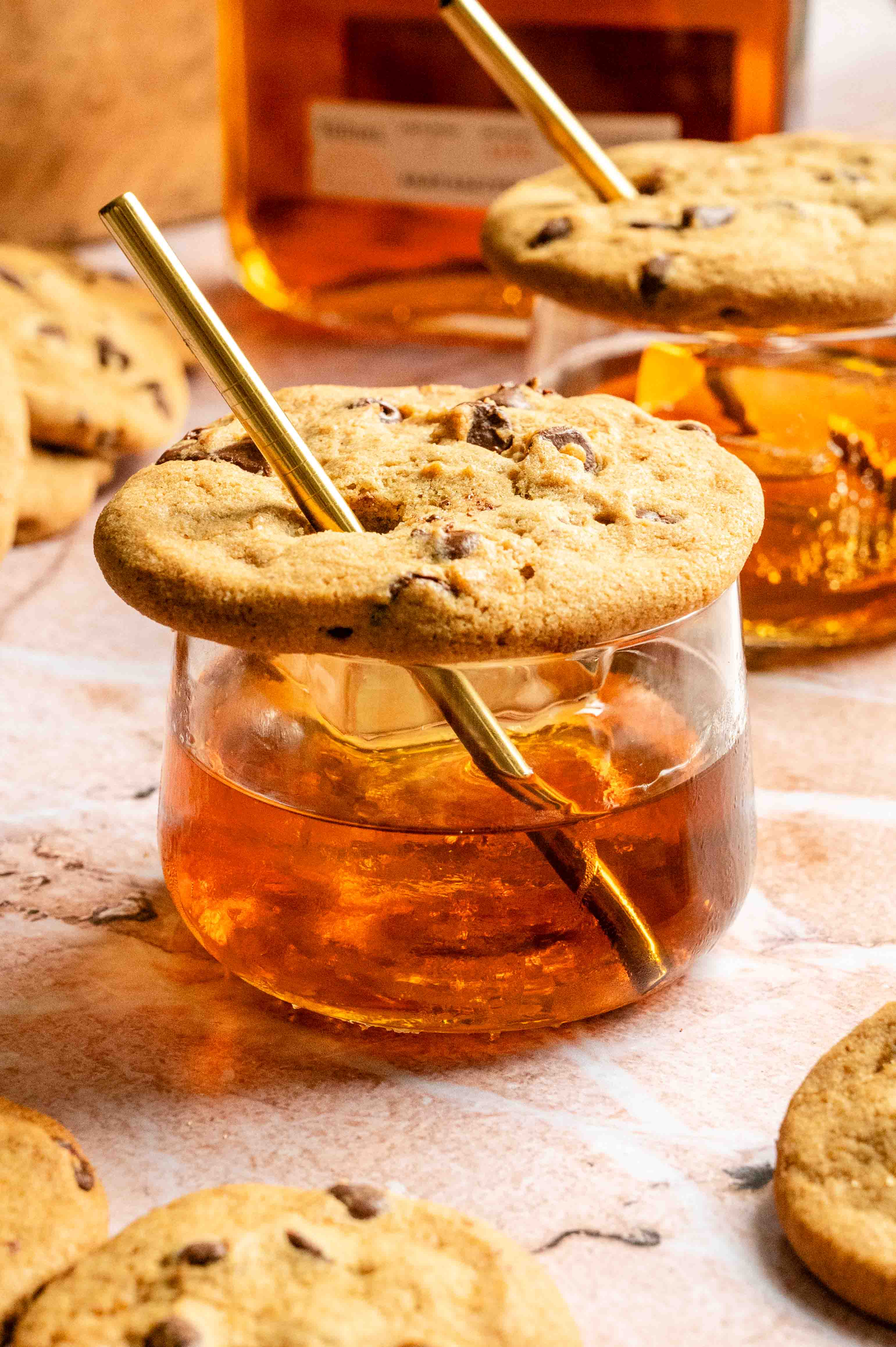 A small old fashioned glass filled with an chocolate chip cookie old fashioned and topped with a chocolate chip cookie with a straw through it.