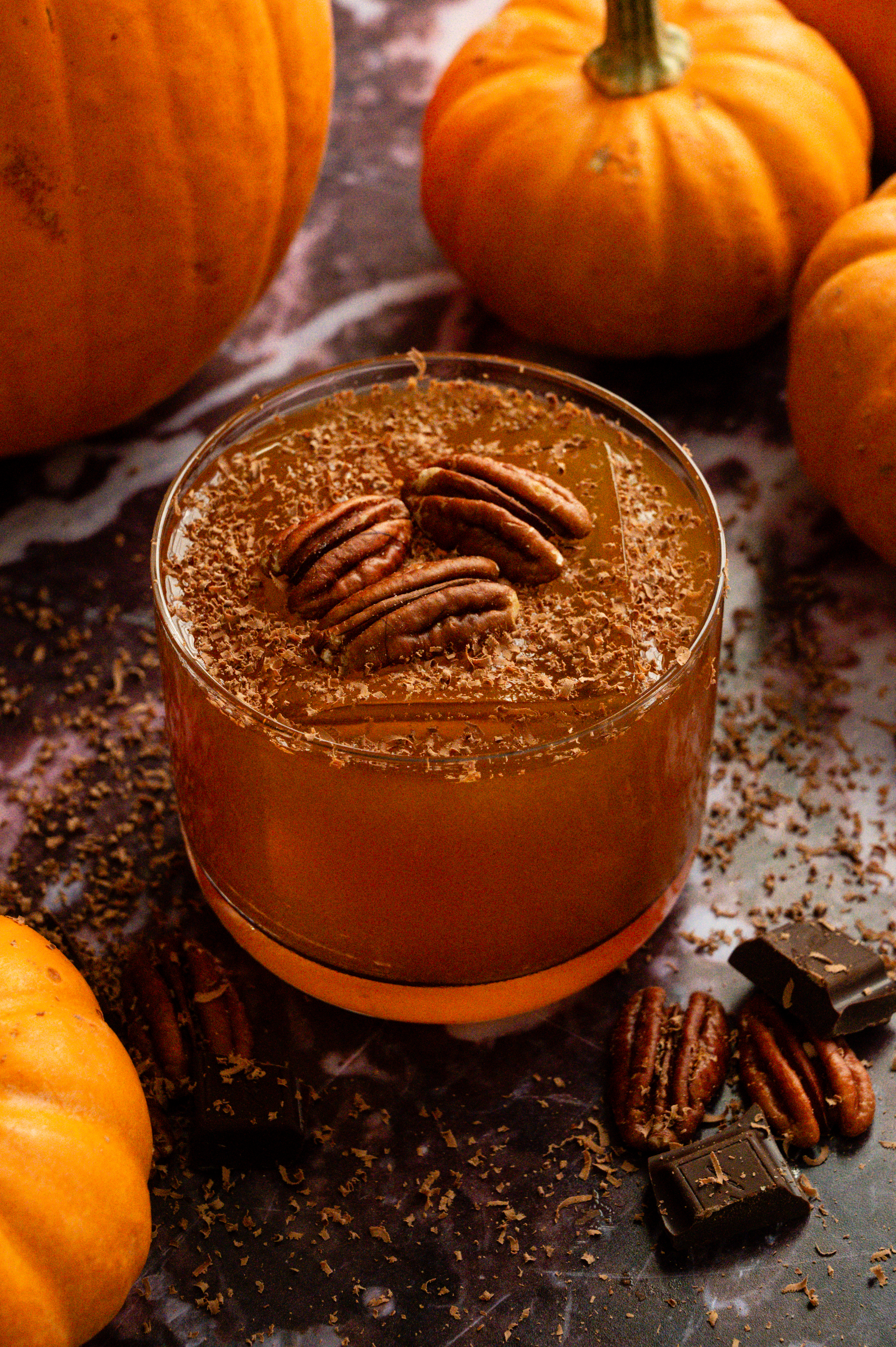 A spiced pumpkin and pecan old fashioned garnished with shaved chocolate and 3 pecans on a purple marble table.