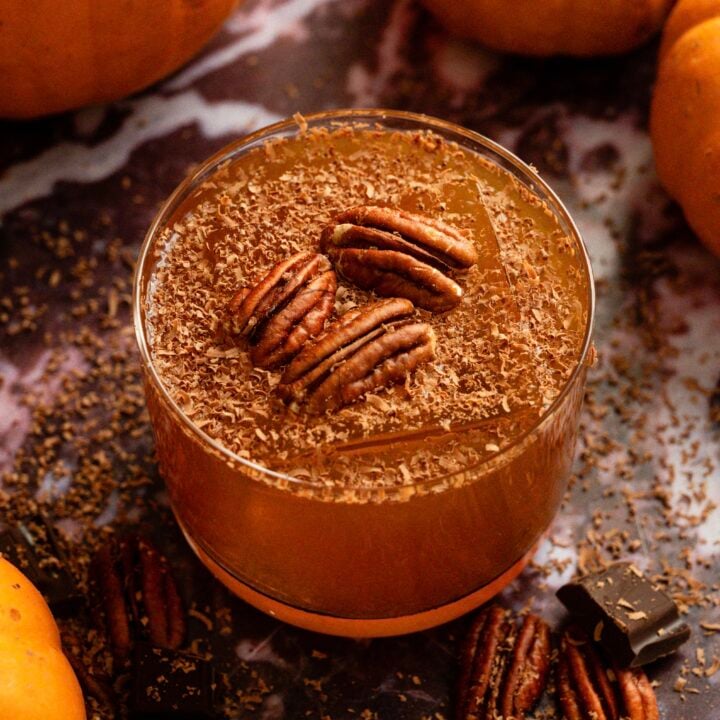 A spiced pumpkin and pecan old fashioned garnished with shaved chocolate and 3 pecans on a purple marble table.