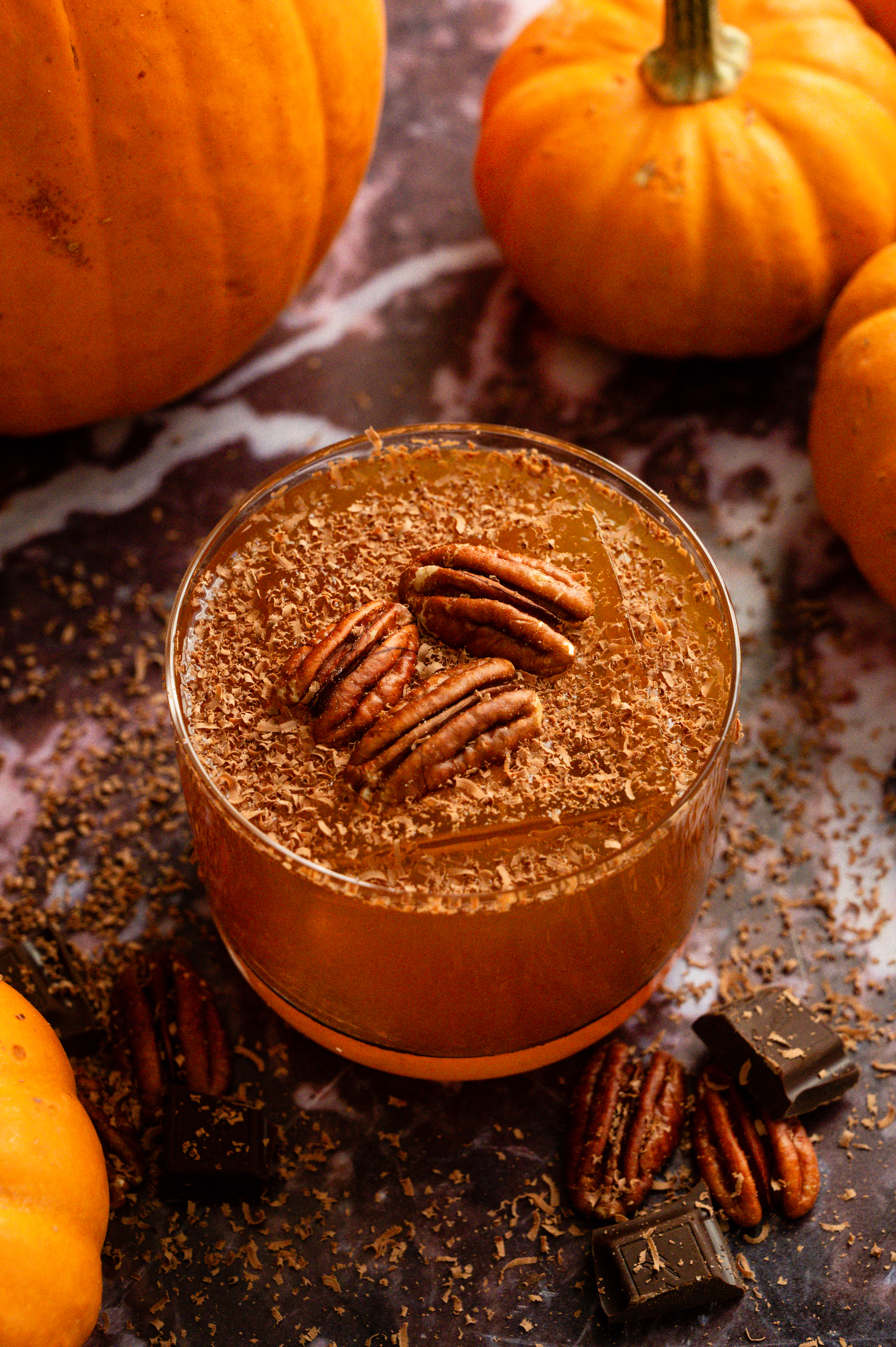 A spiced pumpkin and pecan old fashioned garnished with shaved chocolate and 3 pecans on a purple marble table.